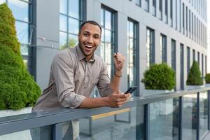 Portrait of successful businessman outside office building, hispanic man smiling and looking at camera celebrating victory and successful achievement holding phone and hand up triumph gesture. photo