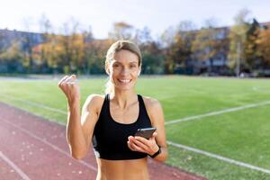 joven hembra atleta comprobación hora en teléfono después correr, maratón. muestra un victoria mano gesto a el cámara. foto