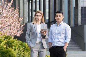 Portrait of two interracial businessmen. A young woman and a young Asian man are standing near the office in suit, Looking at the camera. A woman holds a tablet. A man holds his hands in his pockets. photo