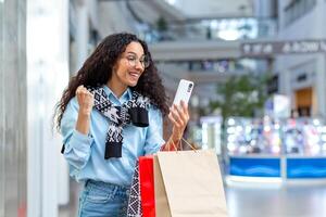 retrato de contento mujer comprador, Hispano mujer dentro centro comercial, usos teléfono inteligente, navega en línea descuentos y ventas, sostiene mano arriba, celebra recibido oferta, ganar foto