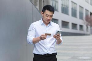 retrato de un joven asiático hombre en pie en el calle cerca un moderno oficina centro, participación un teléfono y un crédito tarjeta en su manos, entrando el tarjeta número, haciendo en línea compras, colocación un orden. foto