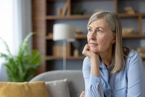 A serene mature woman in a blue shirt stares thoughtfully while sitting in a well-lit living room. The peaceful atmosphere highlights her pensive mood. photo
