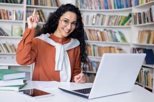 Delighted university student making eureka gesture while sitting by table with pc on background of bookshelves. Intelligent female doing homework and getting right answer for mathematics task. photo