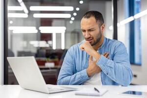 Young hispanic man working and studying in the office, sitting at a desk with a laptop and coughing, holding his hand to his chest and feeling severe pain. photo