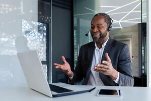 Mature experienced man with headset phone inside office at workplace, smiling talking remotely and advising customers, african american customer support worker, using call and laptop. photo