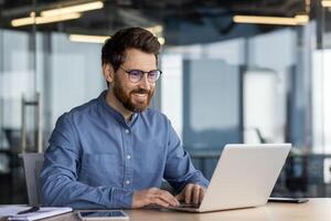 Happy male professional using laptop at desk with bright office background, exuding confidence and productivity. photo