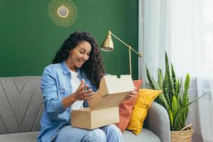 un joven latín americano mujer recibido un agradable paquete o empaquetar entregado por mensajero a su hogar. ella se sienta felizmente en el sofá y desempaqueta el caja. foto