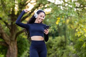 Rejoiced slim woman with long hair raising hand while looking at smartphone screen and wearing headset. Motivated female in black activewear finishing running and ready for taking part in marathon. photo