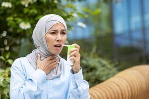 Diverse female with medical aerosol having pulmonary disease and using treatment for relieving bronchial spasms. Stressed lady wearing hijab and struggling with breath disability during stroll. photo