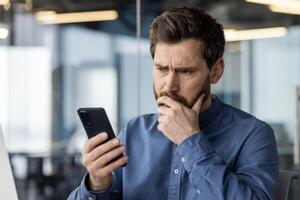 un enfocado masculino adulto con un barba mira a su teléfono inteligente con un preocupado expresión, posiblemente relación comercial con urgente negocio asuntos o malo noticias. foto