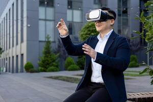 Smiling young man sitting outside office center on bench wearing virtual glasses, gesturing with hands and working remotely, having fun on work break. photo