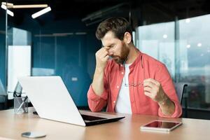 Sick man at workplace has eye pain and dizziness, businessman overworked and overtired working late with laptop sitting at desk inside office. photo