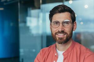 Close-up photo of happy and successful smiling programmer in red casual shirt, man in glasses looking at camera and smiling, developer engineer working inside office.