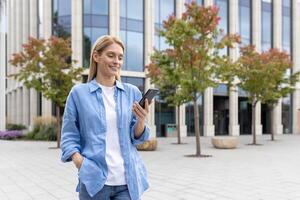 Mature woman with phone in hands walking in the city, a businesswoman in blue shirt holding smartphone in hands, reading online social networks, blonde smiling satisfaction is using an application. photo