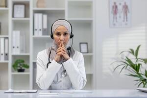 Focused female doctor wearing a hijab engaged in a serious call, looking directly at the camera in a modern office setting. photo