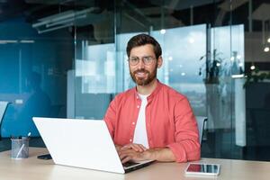 Portrait of mature businessman freelancer startup, bearded man smiling and looking at camera, business owner working inside modern office building wearing red shirt and glasses. photo