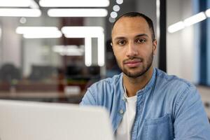 Focused professional man using a laptop in a well-lit contemporary office. The image conveys concepts of business, productivity, tech-savviness, and career-focused determination in a corporate environment. photo