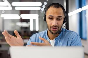 Close-up photo of a smiling young hispanic man in a headset sitting in the office in front of a laptop and talking on a call ,while gesturing with his hands.