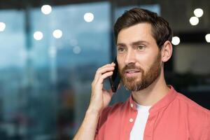 Successful businessman in a red shirt talking on the phone inside the office, a man with a beard at the workplace is talking to a client, a satisfied boss in casual clothes. photo