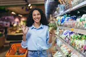 Smiling young beautiful Hispanic woman shopping in a supermarket, holding broccoli in her hand photo