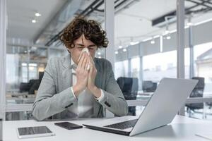 Exhausted young adult businessman caught a cold, working with a laptop at the office and sneezing into a tissue. Displaying common office work fatigue. photo