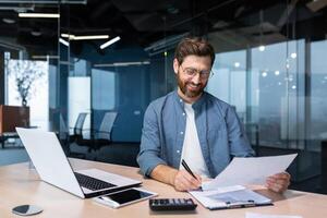 Mature businessman in shirt doing paperwork, man working with documents, contracts and bills sitting at table using laptop at work, financier accountant with beard and glasses. photo