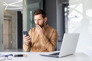 Man reading sad bad news using phone, businessman at workplace holding smartphone, shocked and disappointed inside office with laptop. photo