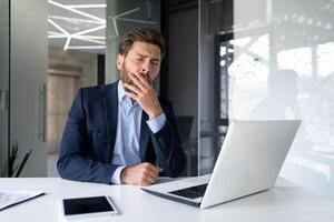 Tired young businessman man in a suit sits in the office at the desk and yawns, covers his mouth with his hand, feels sleepy. photo