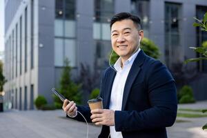 Portrait of an Asian young male office worker wearing headphones and holding a phone and a cup of coffee resting sitting outside an office ,center and smiling at the camera. photo