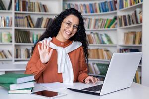 un joven hembra alumno, rodeado por libros, olas a su ordenador portátil pantalla en un biblioteca configuración, sugerencia un virtual Hola. foto