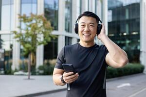 Close-up portrait of a young Asian male athlete standing on a city street wearing headphones, holding a phone in his hand and looking at the camera smiling. photo