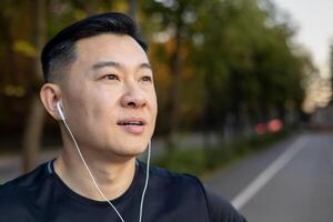 A close-up photo of a young serious man Asian male sportsman standing on the road in the middle of the street in headphones, listening to music, resting, looking to the side.