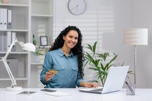 exitoso Hispano mujer de negocios utilizando crédito tarjeta para en línea compras mientras trabajando desde el comodidad de su hogar oficina. foto