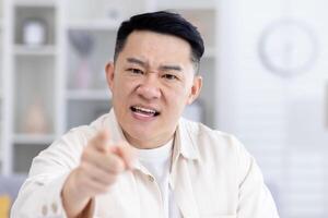 call online meeting, mature adult asian looking at camera angry man shouting angry, businessman working remotely from home talking to colleagues, boss nervous at home talking to subordinates. photo