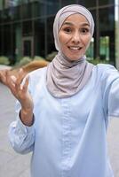 un vibrante Disparo de un joven musulmán mujer en un azul hijab capturar un selfie con un moderno urbano fondo. foto