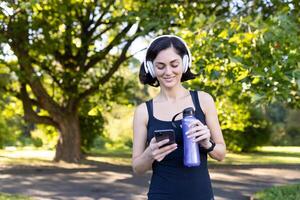 Smiling young woman doing sports and jogging outdoors in park, standing resting in headphones, holding water bottle and looking at phone screen. photo