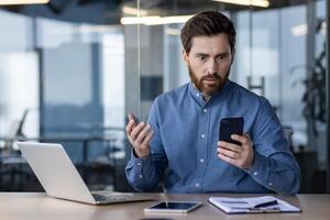 Bearded businessman in blue shirt looks shocked while reading a message on his smartphone with laptop and tablet on desk in a modern office. photo