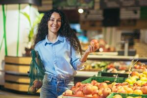 Portrait of happy and smiling hispanic female shopper in supermarket, woman smiling and looking at camera chooses apples and puts in eco bag photo