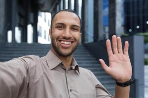 Young african american man looking into smartphone camera, talking on call using app on phone, smiling and happy waving hand gesture of greeting, businessman outside modern office building photo