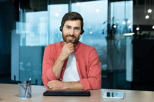 call, joyful smiling man with headset phone working in evening time inside office, web camera view, online meeting with colleagues partners remotely. photo