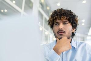 un joven pelo rizado hombre vestido en un azul camisa, reflexionando profundamente mientras sentado en un brillante, contemporáneo oficina ambiente. su expresión refleja concentración y contemplación. foto