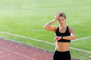 cansado y trastornado maduro hembra atleta comprobación corazón Velocidad en inteligente reloj aptitud pulsera, corredor en estadio después activo ejercicio y trotar en soleado día, rubia mujer en ropa de deporte. foto