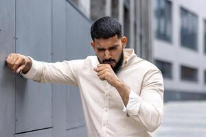 Cough attack. A young Indian man is standing outside the building, holding on to the wall with his hand, covering his mouth with his hand and coughing, breathing hard. Close-up photo