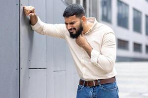 A young Indian man is standing outside, leaning against the wall of the building and holding his hand around his neck, feeling severe pain and discomfort in his throat photo