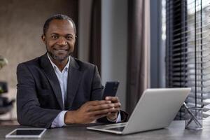 retrato de alegre maduro africano americano jefe en negocio traje, mayor empresario sonriente y mirando a cámara con teléfono en manos, hombre trabajando dentro oficina con computadora portátil, utilizando aplicación foto