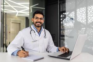 Confident male doctor with stethoscope working on laptop in modern office photo