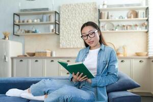 joven hermosa asiático mujer en lentes y mezclilla camisa estudiando a hogar, contento hembra estudiante sonriente leyendo libro sentado en sofá en vivo habitación foto