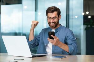 empresario celebrando éxito y triunfo, hombre en camisa trabajando dentro moderno oficina con ordenador portátil participación teléfono leyendo mensaje, negocio propietario recibido bueno Noticias notificación en línea foto