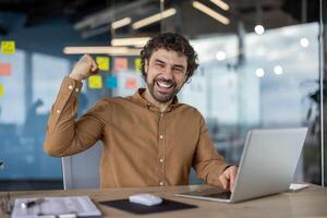 un alegre empresario con Rizado pelo triunfantemente celebrando un éxito en un oficina ajuste con un ordenador portátil en frente de a él. foto