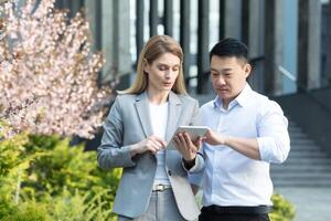 Interracial partnership. Young business woman and young Asian business man standing near office center, talking, working on tablet, discussing business, cooperation, agreement. photo